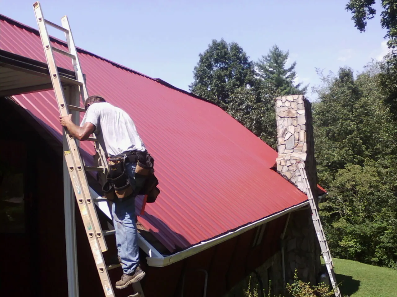 Roofer on a ladder beside a red metal roof and stone chimney — Soffit & Fascia Repair by Roofing Brothers in Sapphire